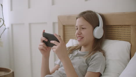 cheerful-little-girl-is-lying-on-bed-with-smartphone-in-hands-and-playing-game-spend-time-in-internet